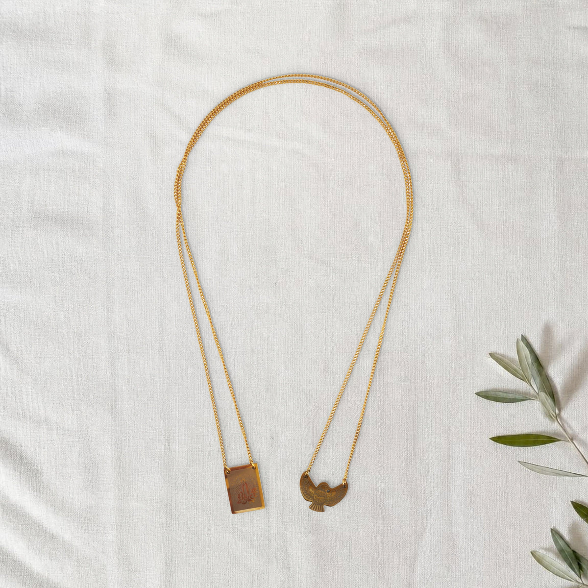 Colar Escapulário Anunciação dourado em cenário de linho com raminho de oliveira.