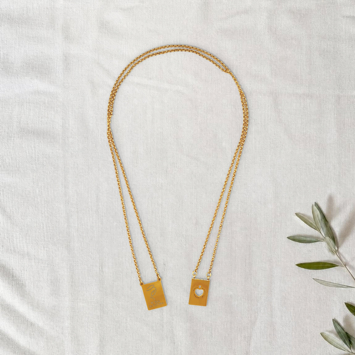 Colar Escapulário Refúgio dourado em cenário de linho com raminho de oliveira.