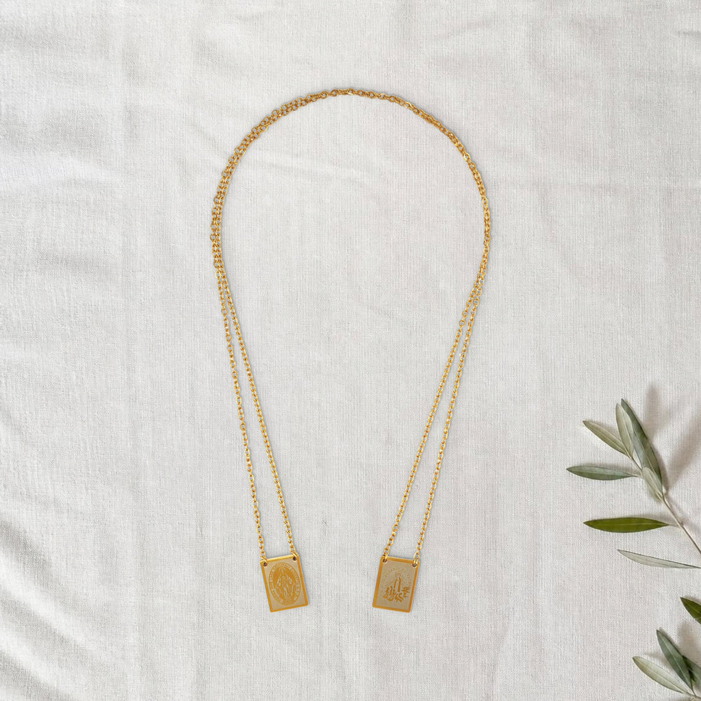 Colar Escapulário Candeia dourado em cenário de linho com raminho de oliveira.