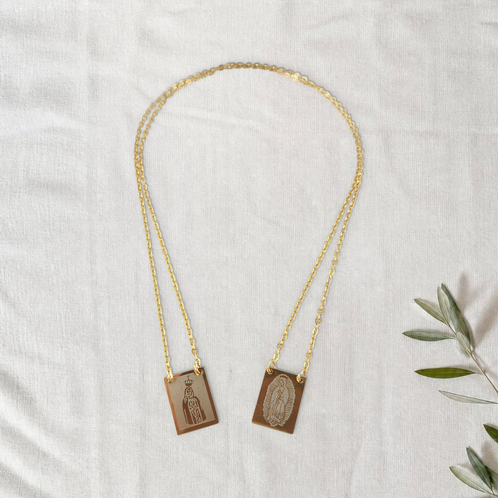 Colar Escapulário Além-Mar dourado em cenário de linho com raminho de oliveira.