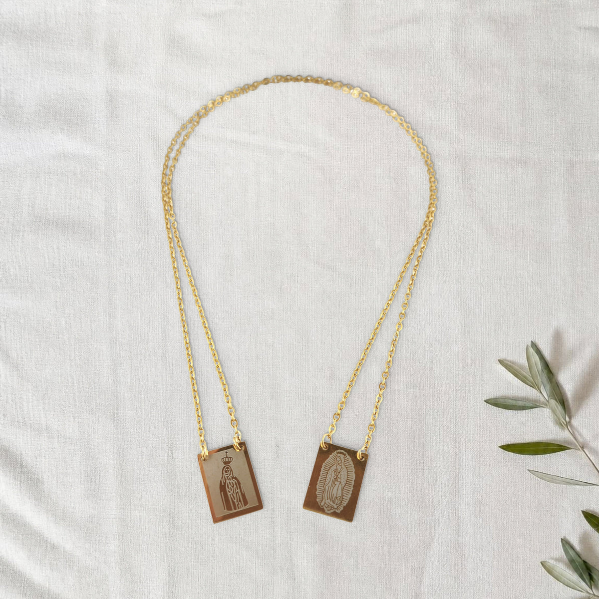 Colar Escapulário Além-Mar dourado em cenário de linho com raminho de oliveira.
