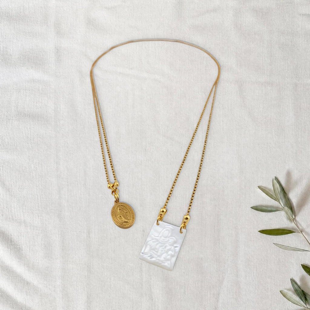 Colar Escapulário Manto de Graça dourado em cenário de linho com raminho de oliveira.