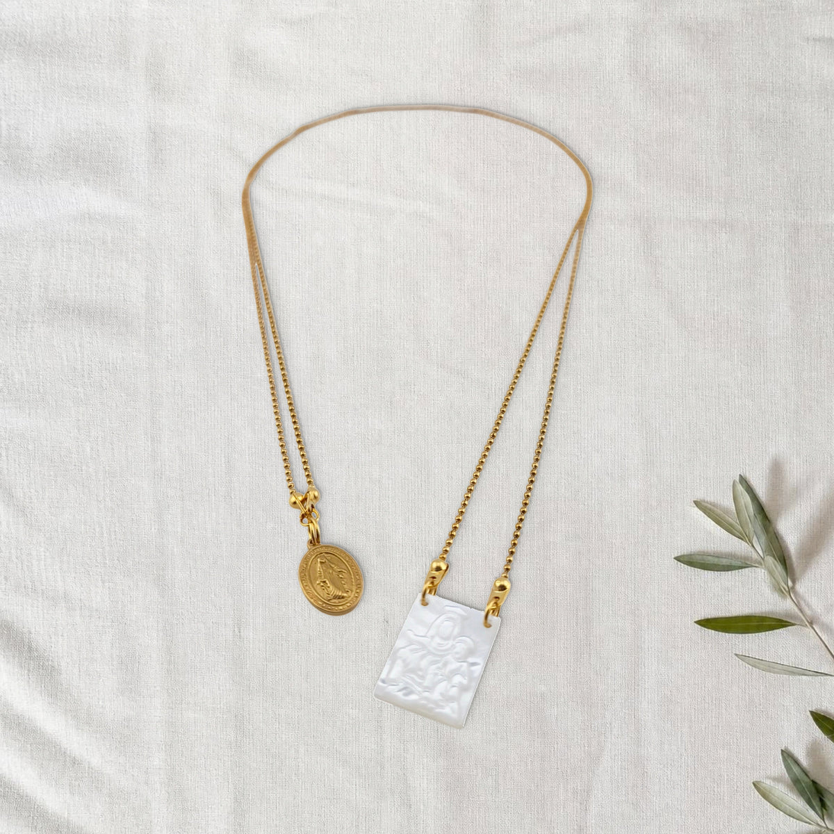 Colar Escapulário Manto de Graça dourado em cenário de linho com raminho de oliveira.