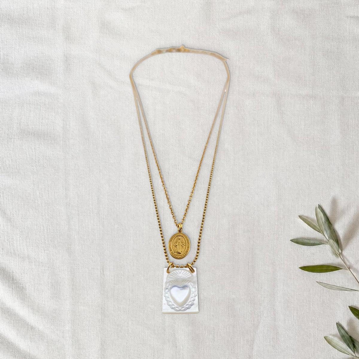 Colar Escapulário Devoção dourado em cenário de linho com raminho de oliveira.