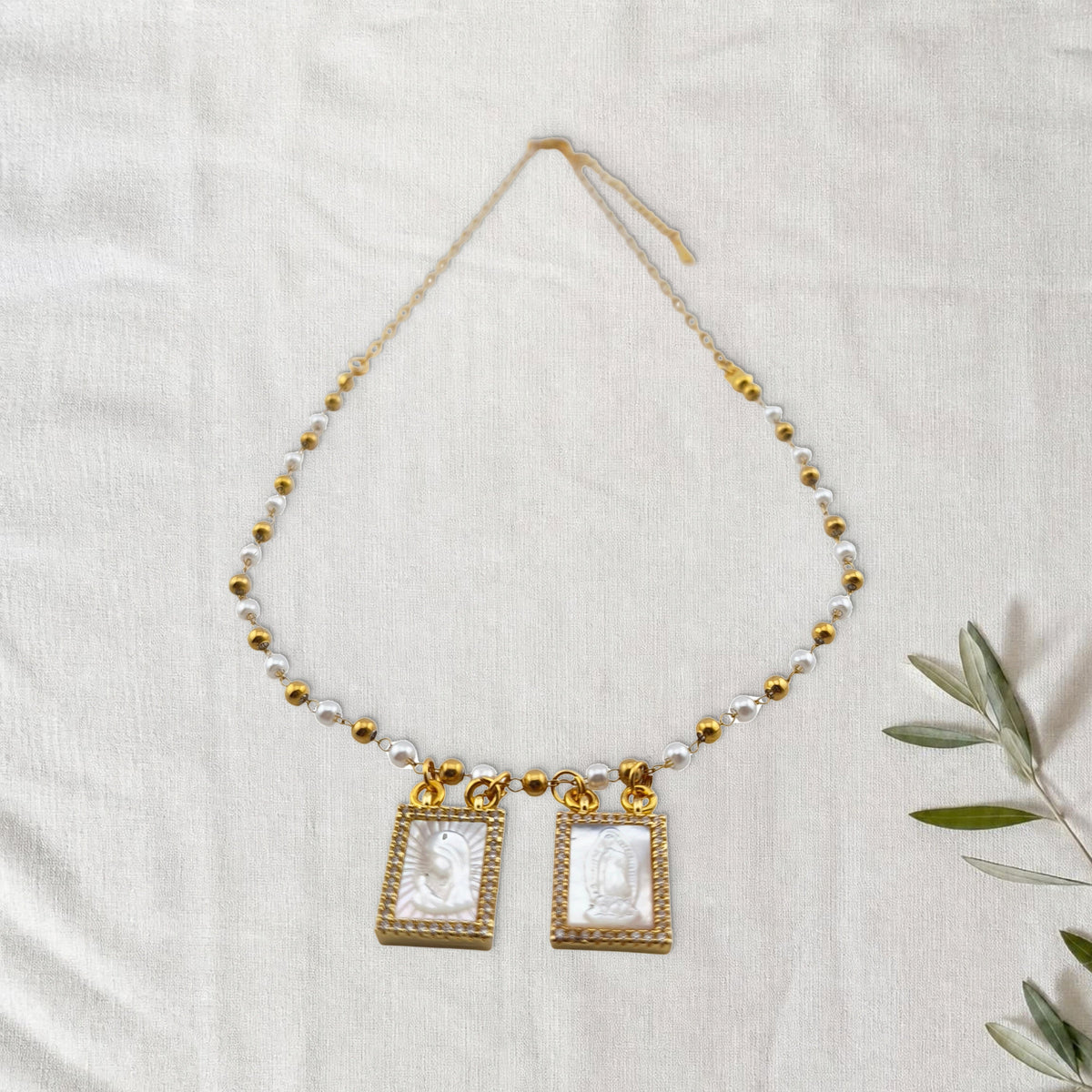 Colar Escapulário Luz Celestial dourado em cenário de linho com raminho de oliveira.
