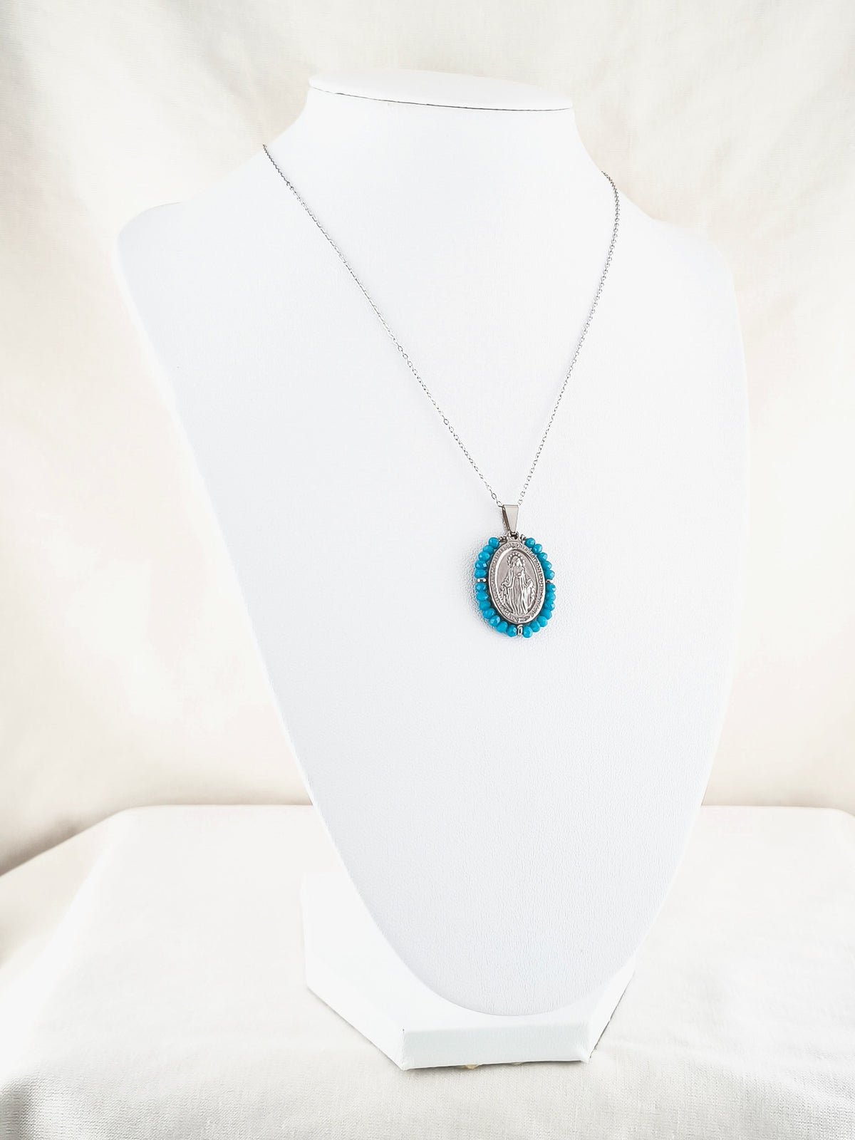 Fio Auréola prateado, com medalha de Nossa Senhora das Graças rodeada de contas turquesa, apresentado em expositor.