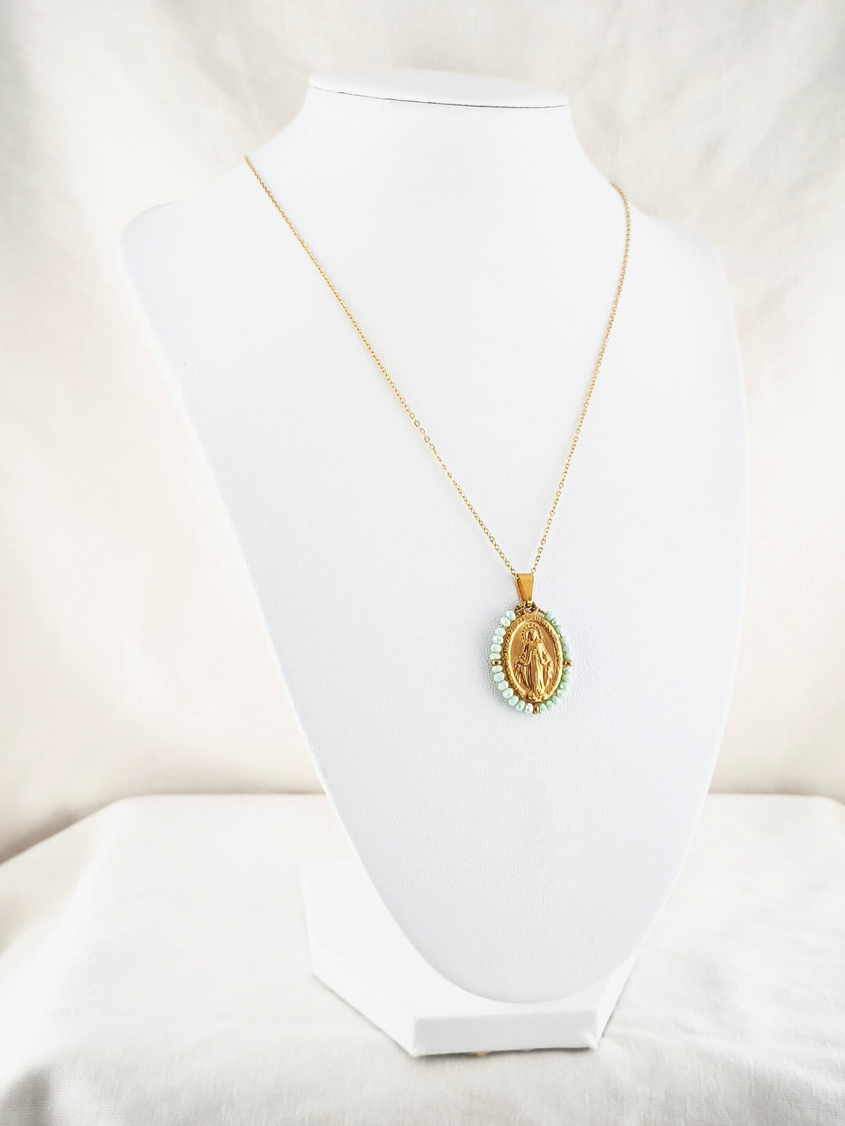 Fio Auréola dourado, com medalha de Nossa Senhora das Graças rodeada de contas verde-água, apresentado em expositor.