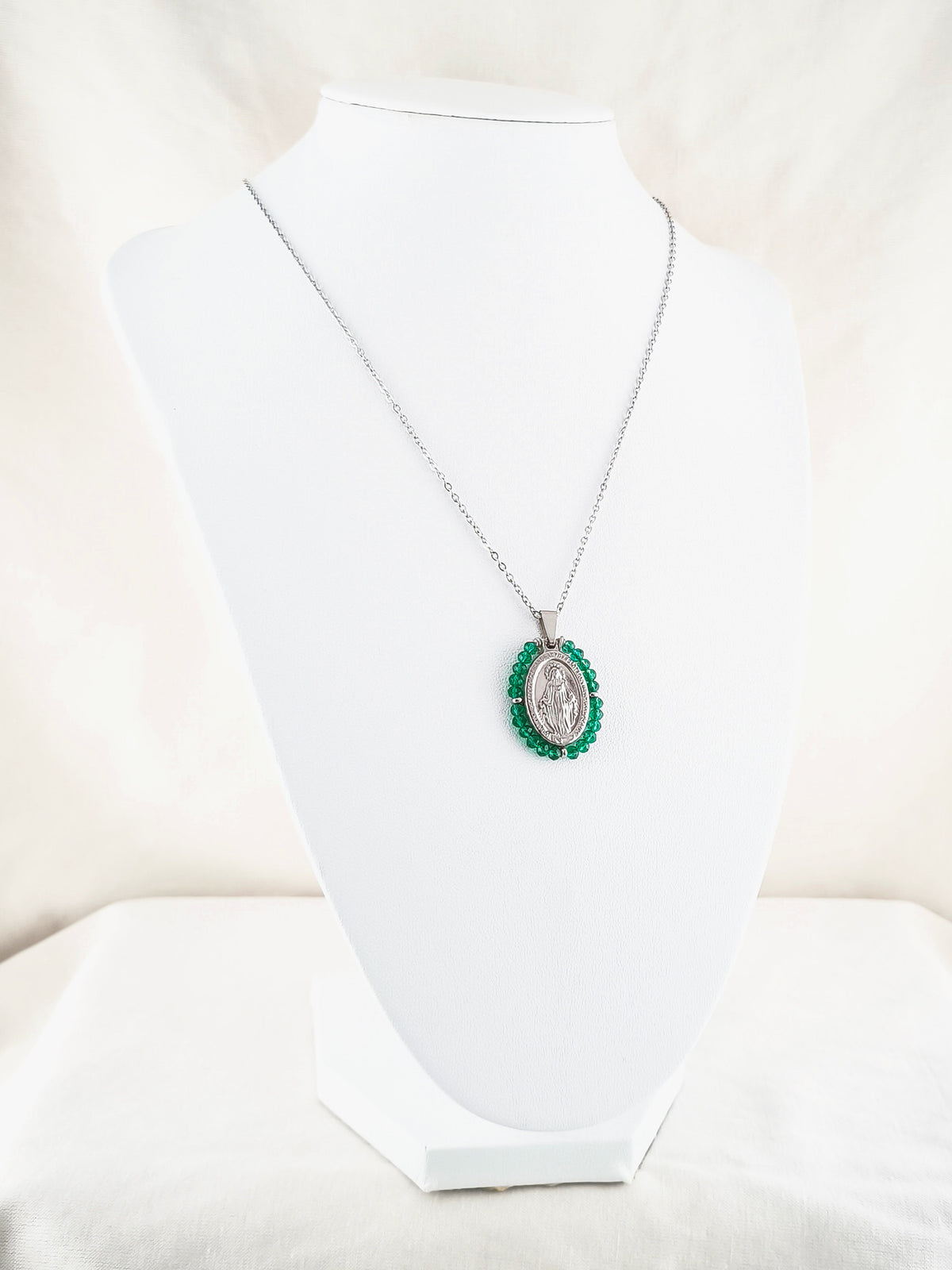 Fio Auréola prateado, com medalha de Nossa Senhora das Graças rodeada de contas verde esmeralda, apresentado em expositor.