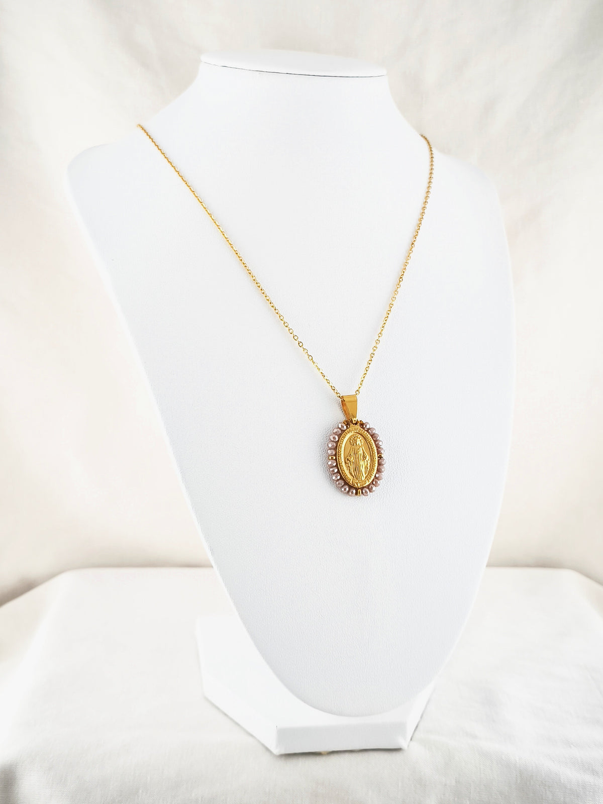 Fio Auréola dourado, com medalha de Nossa Senhora das Graças rodeada de contas rosa irisado, apresentado em expositor.