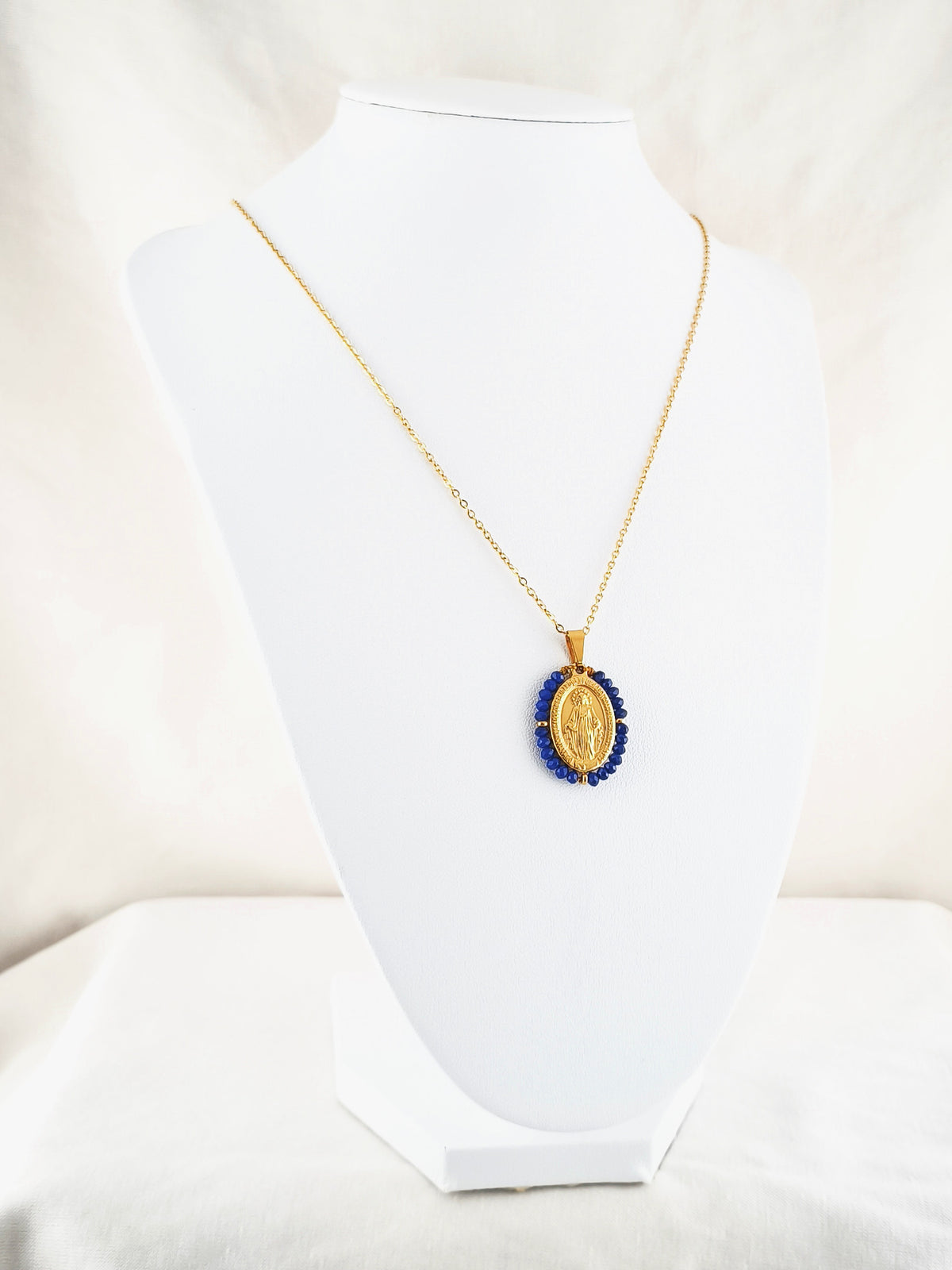 Fio Auréola dourado, com medalha de Nossa Senhora das Graças rodeada de contas azul cobalto, apresentado em expositor.
