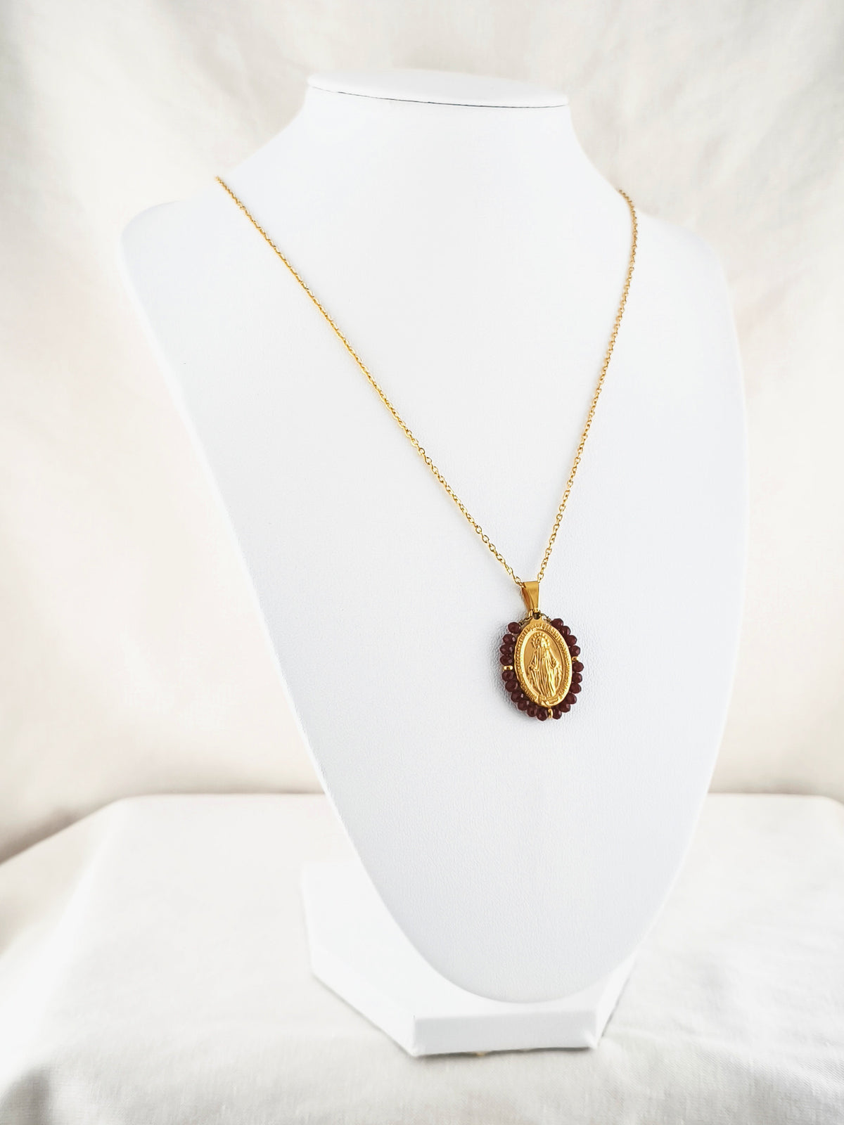 Fio Auréola dourado, com medalha de Nossa Senhora das Graças rodeada de contas vinho, apresentado em expositor.