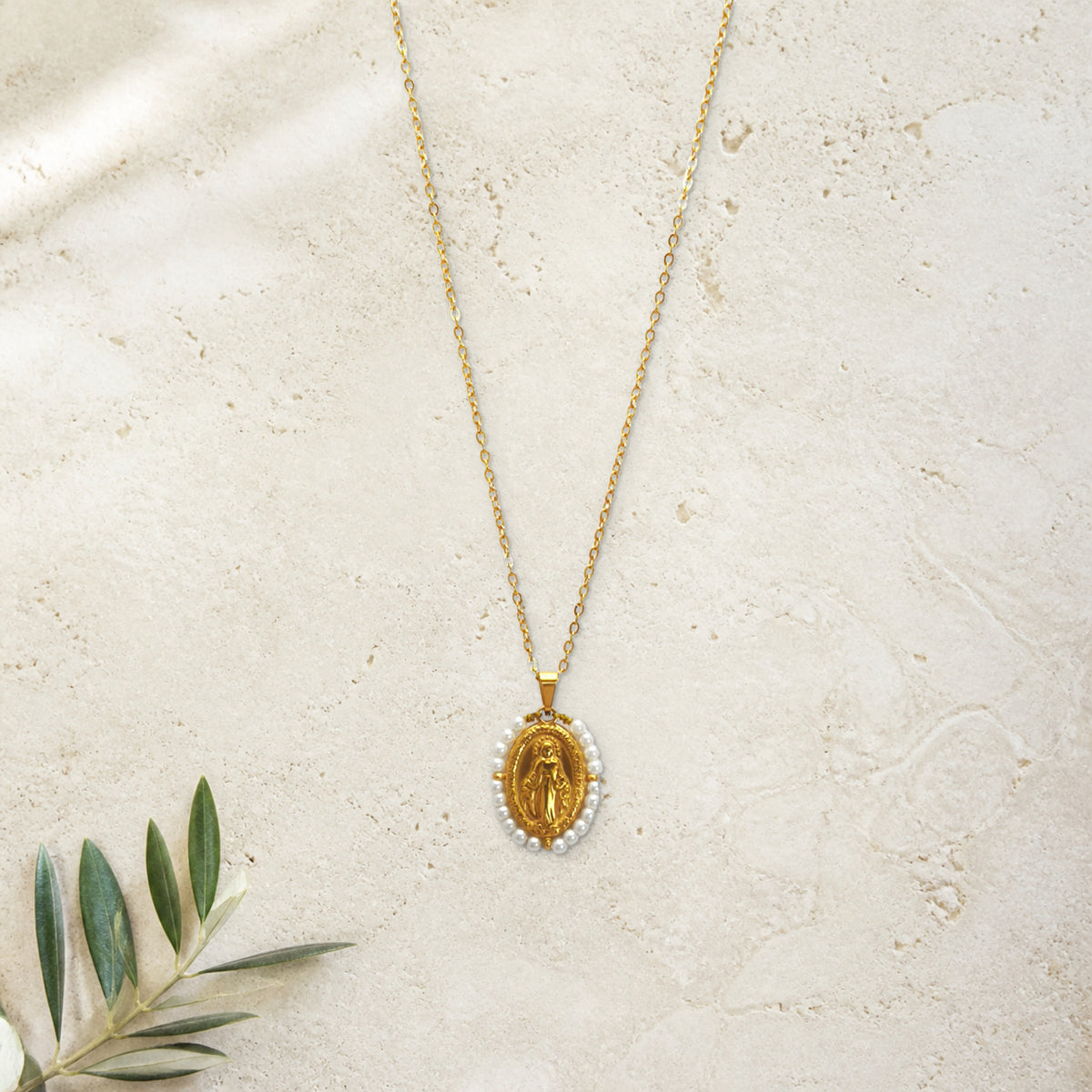 Fio Auréola dourado com pérolas artificiais e medalha de Nossa Senhora das Graças, pousado sobre pedra travertino com um raminho.