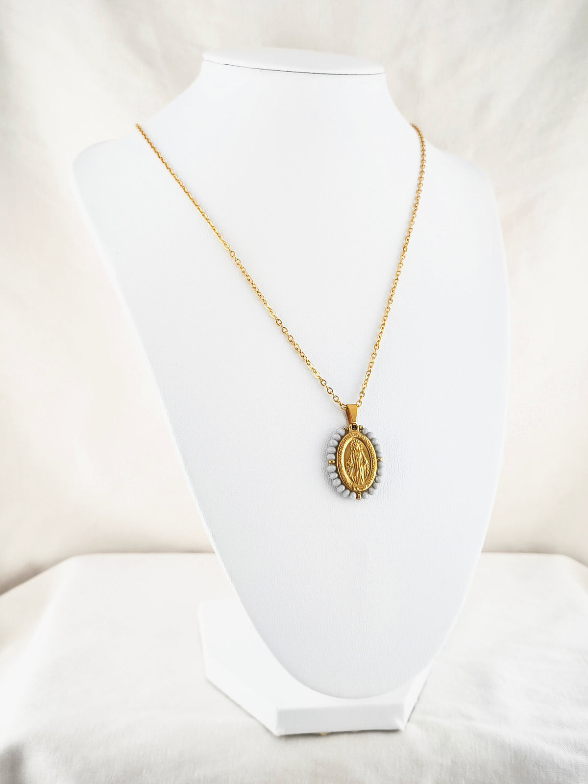 Fio Auréola dourado, com medalha de Nossa Senhora das Graças rodeada de contas cinza claro, apresentado em expositor.