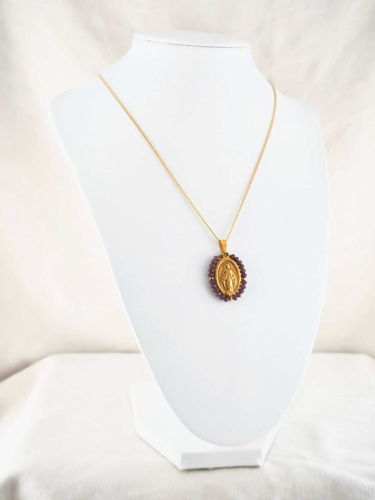 Fio Auréola dourado, com medalha de Nossa Senhora das Graças rodeada de contas ameixa, apresentado em expositor.