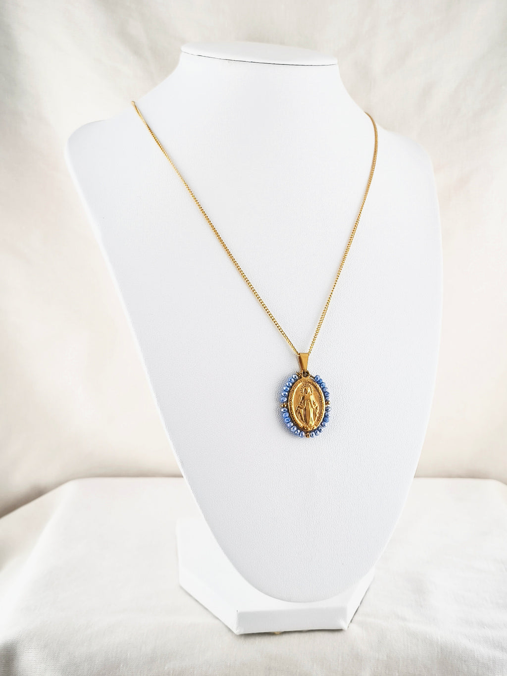 Fio Auréola dourado, com medalha de Nossa Senhora das Graças rodeada de contas azul céu, apresentado em expositor.