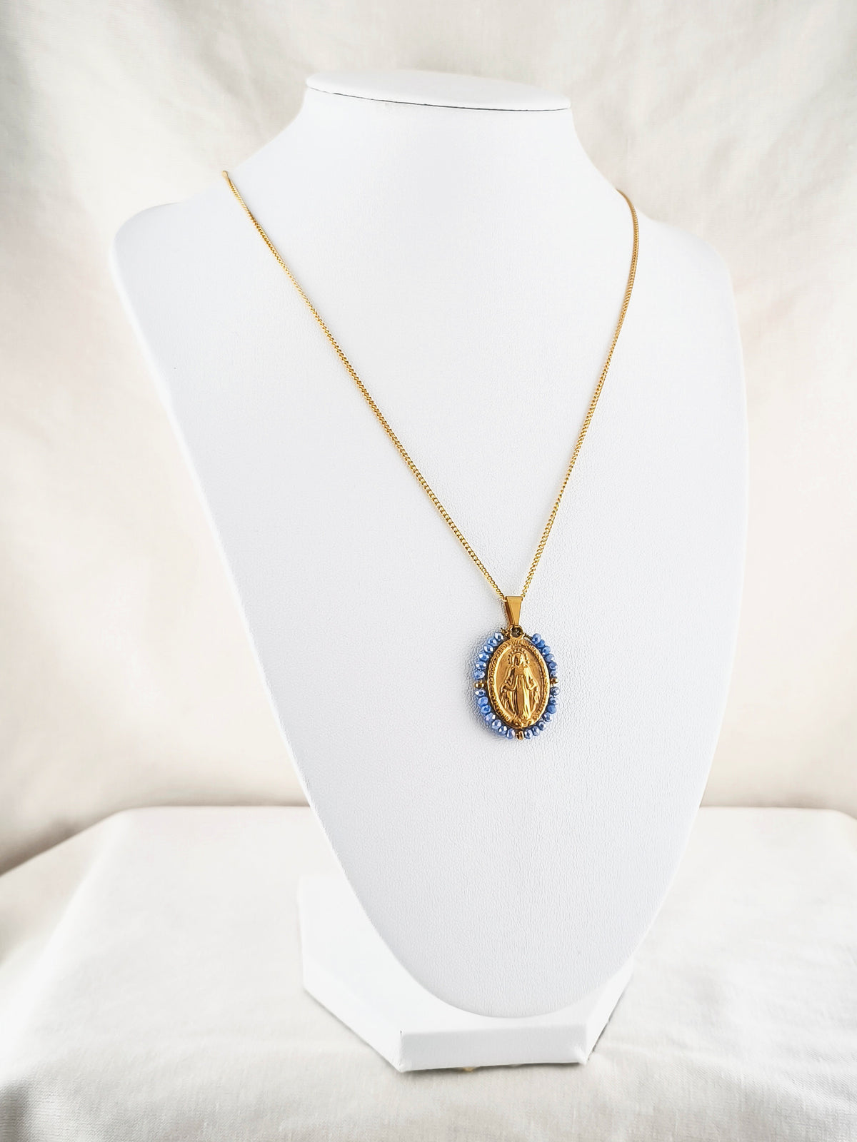 Fio Auréola dourado, com medalha de Nossa Senhora das Graças rodeada de contas azul céu, apresentado em expositor.