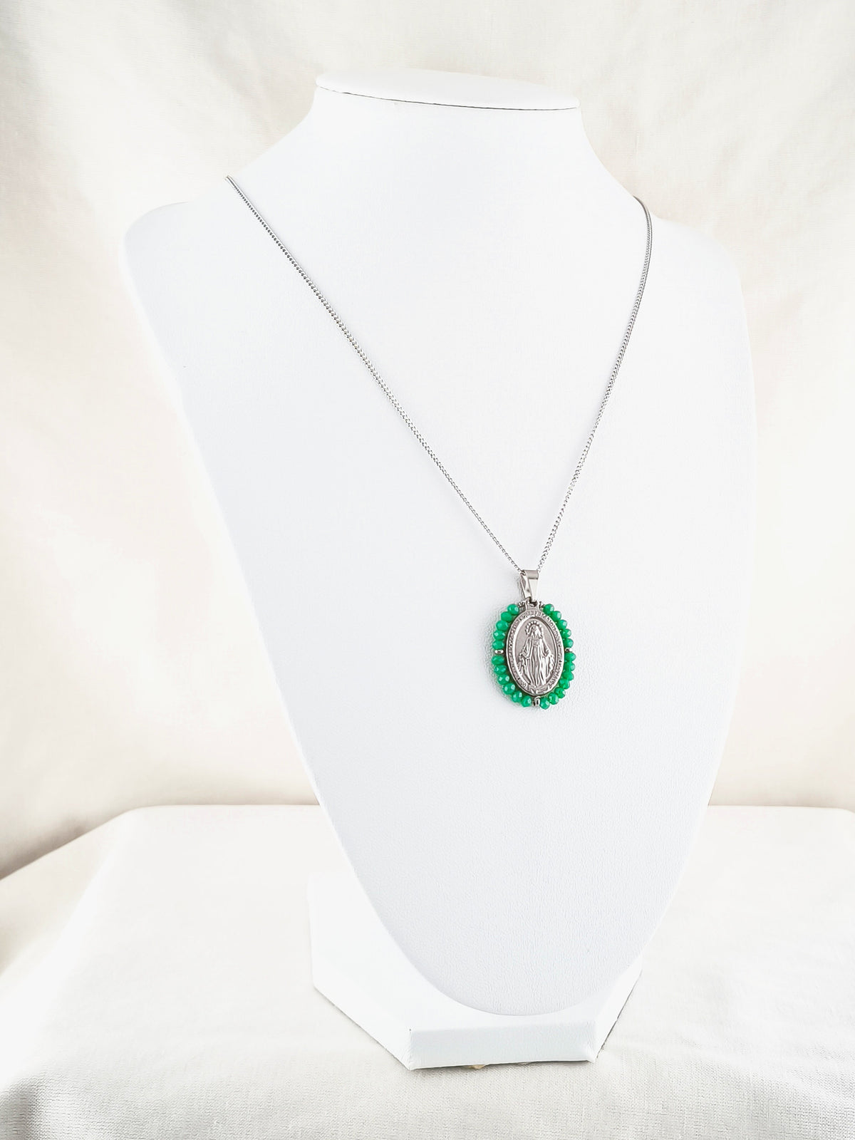 Fio Auréola prateado, com medalha de Nossa Senhora das Graças rodeada de contas verdes, apresentado em expositor.