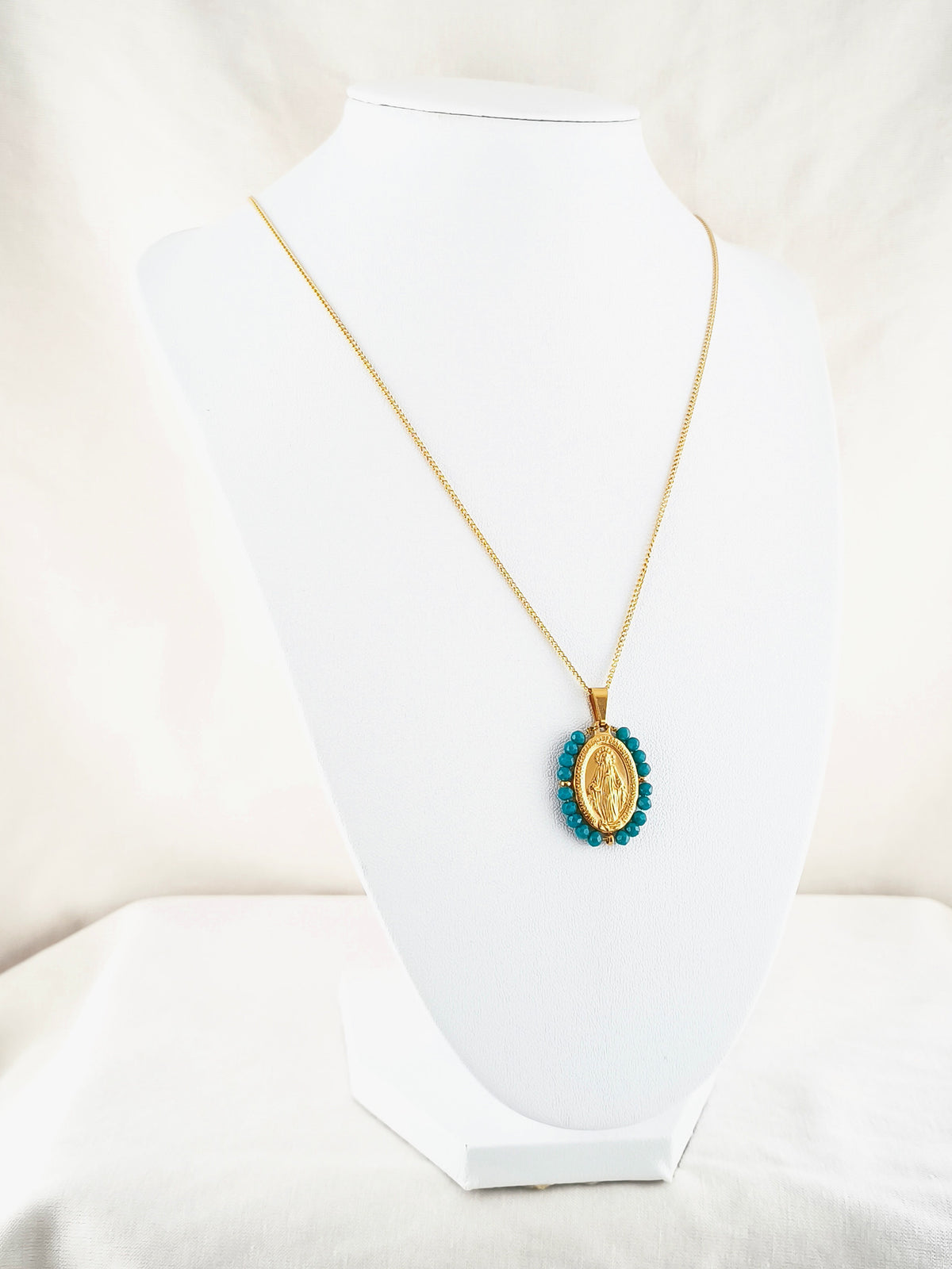 Fio Auréola dourado, com medalha de Nossa Senhora das Graças rodeada de contas turquesa escuro, apresentado em expositor.