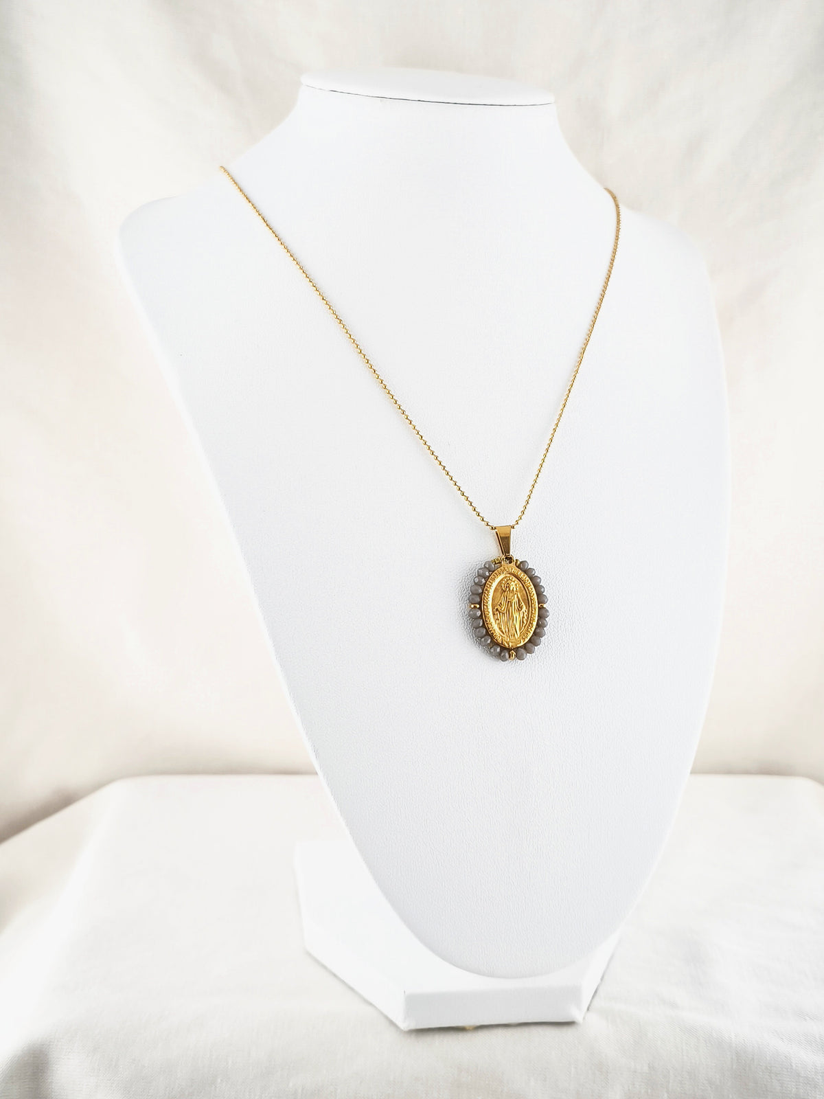 Fio Auréola dourado, com medalha de Nossa Senhora das Graças rodeada de contas cinza, apresentado em expositor.