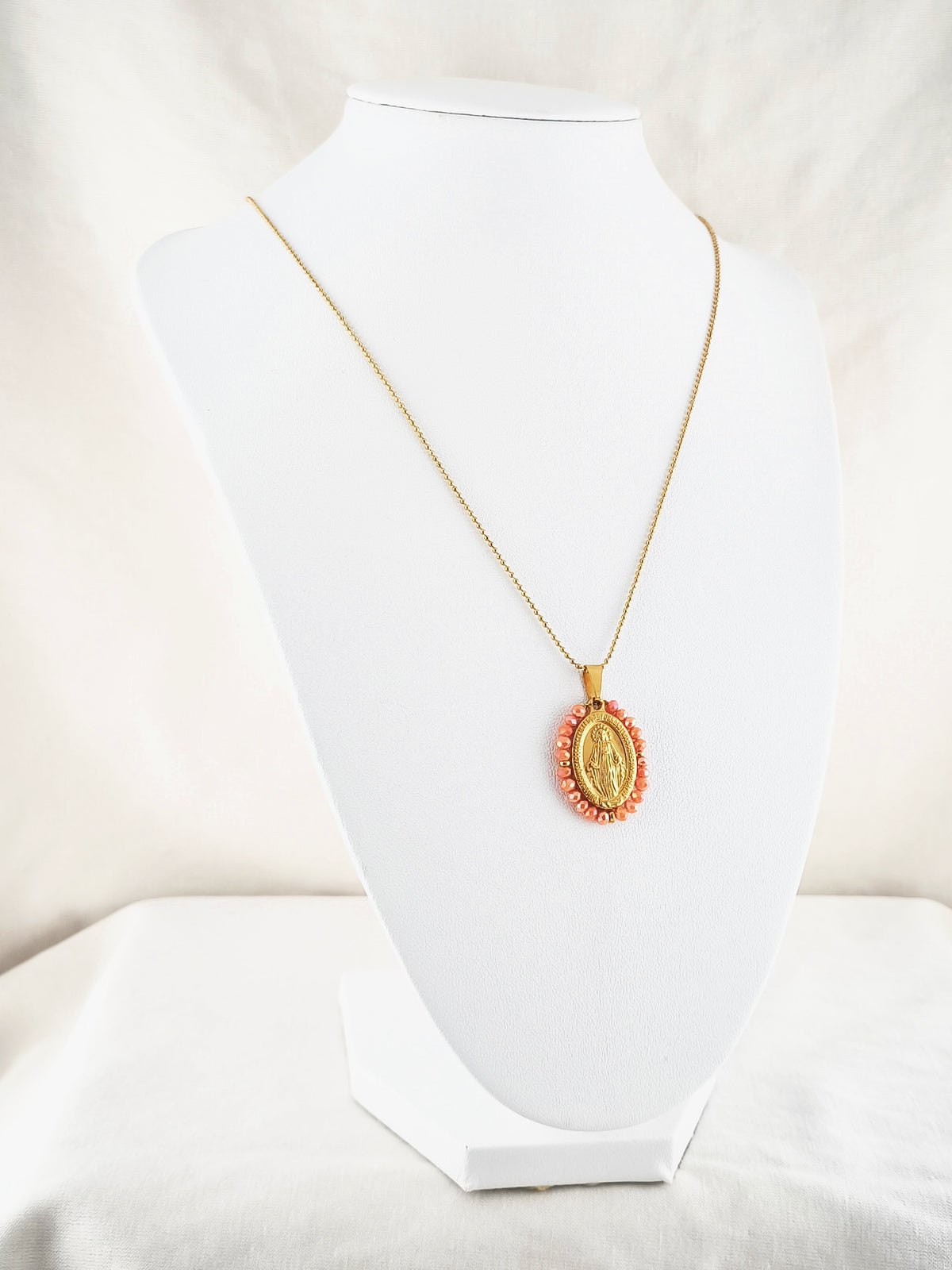 Fio Auréola dourado, com medalha de Nossa Senhora das Graças rodeada de contas salmão, apresentado em expositor.