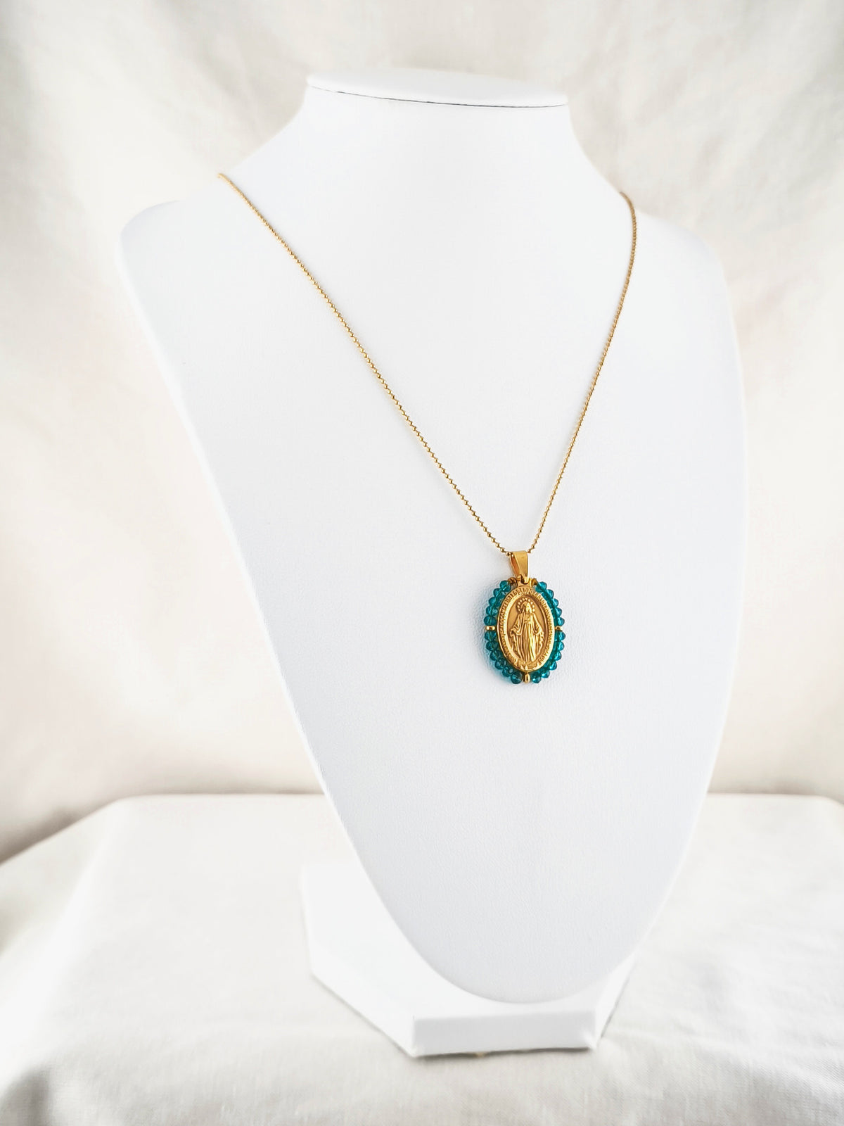 Fio Auréola dourado, com medalha de Nossa Senhora das Graças rodeada de contas petróleo, apresentado em expositor.