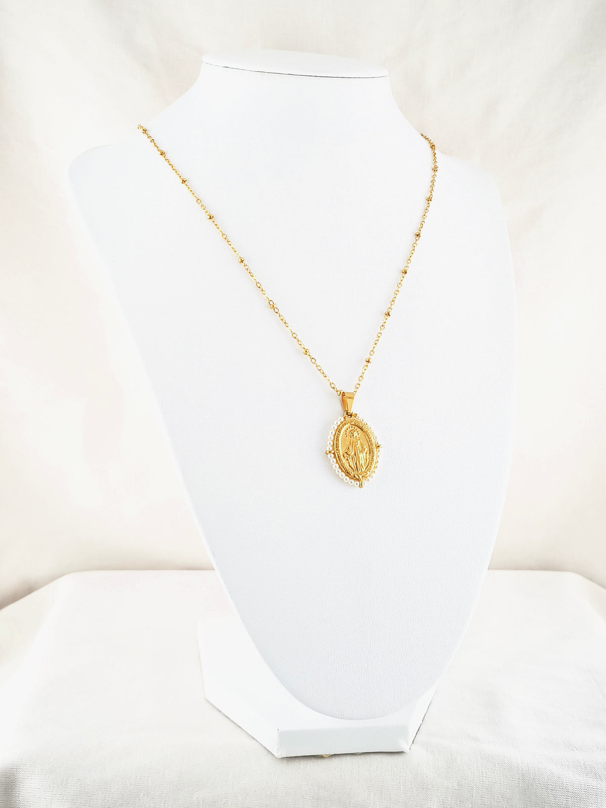 Fio Auréola dourado, com medalha de Nossa Senhora das Graças rodeada de pérolas artificiais miúdas, apresentado em expositor.
