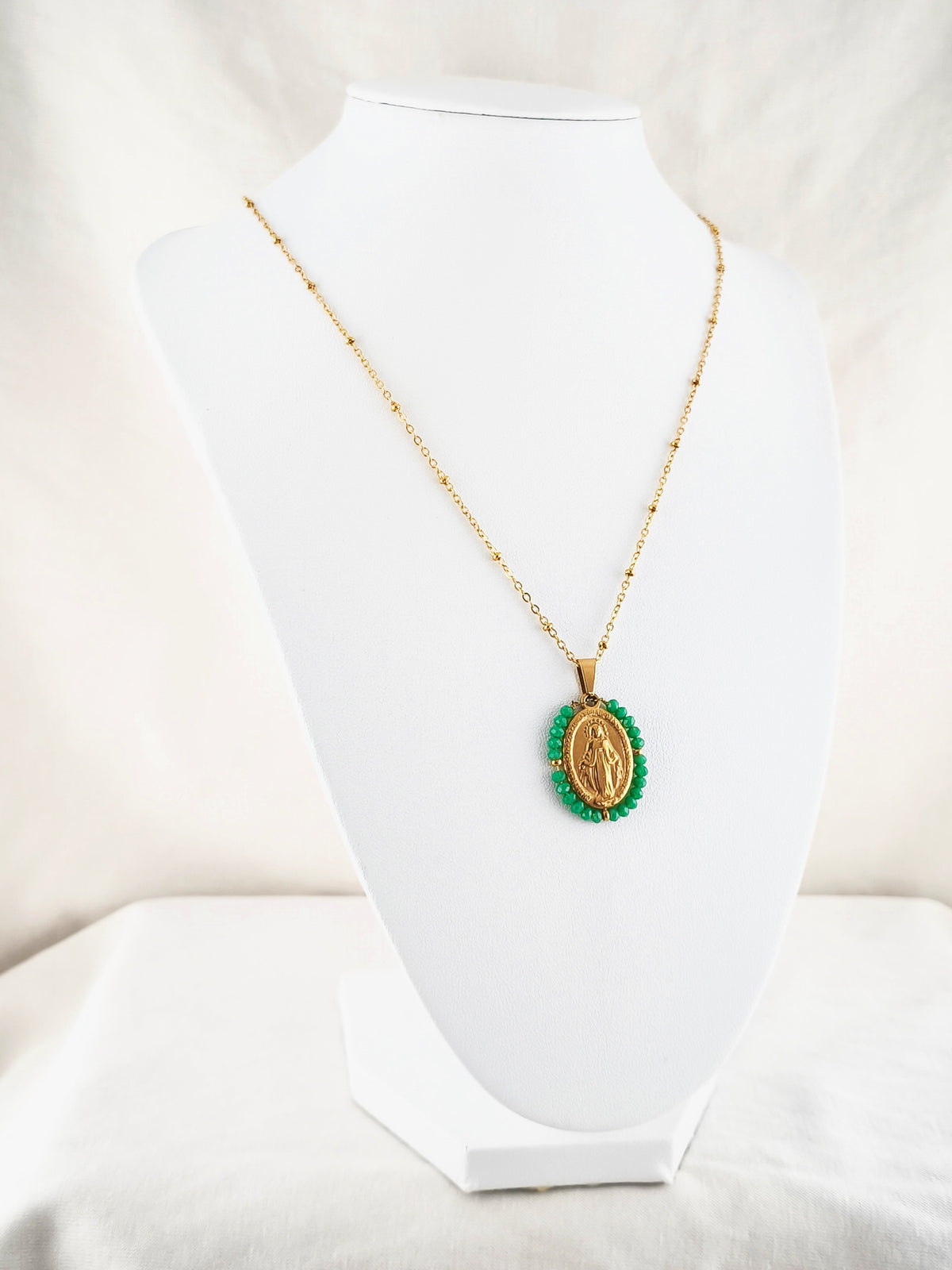 Fio Auréola dourado, com medalha de Nossa Senhora das Graças rodeada de contas verdes, apresentado em expositor.