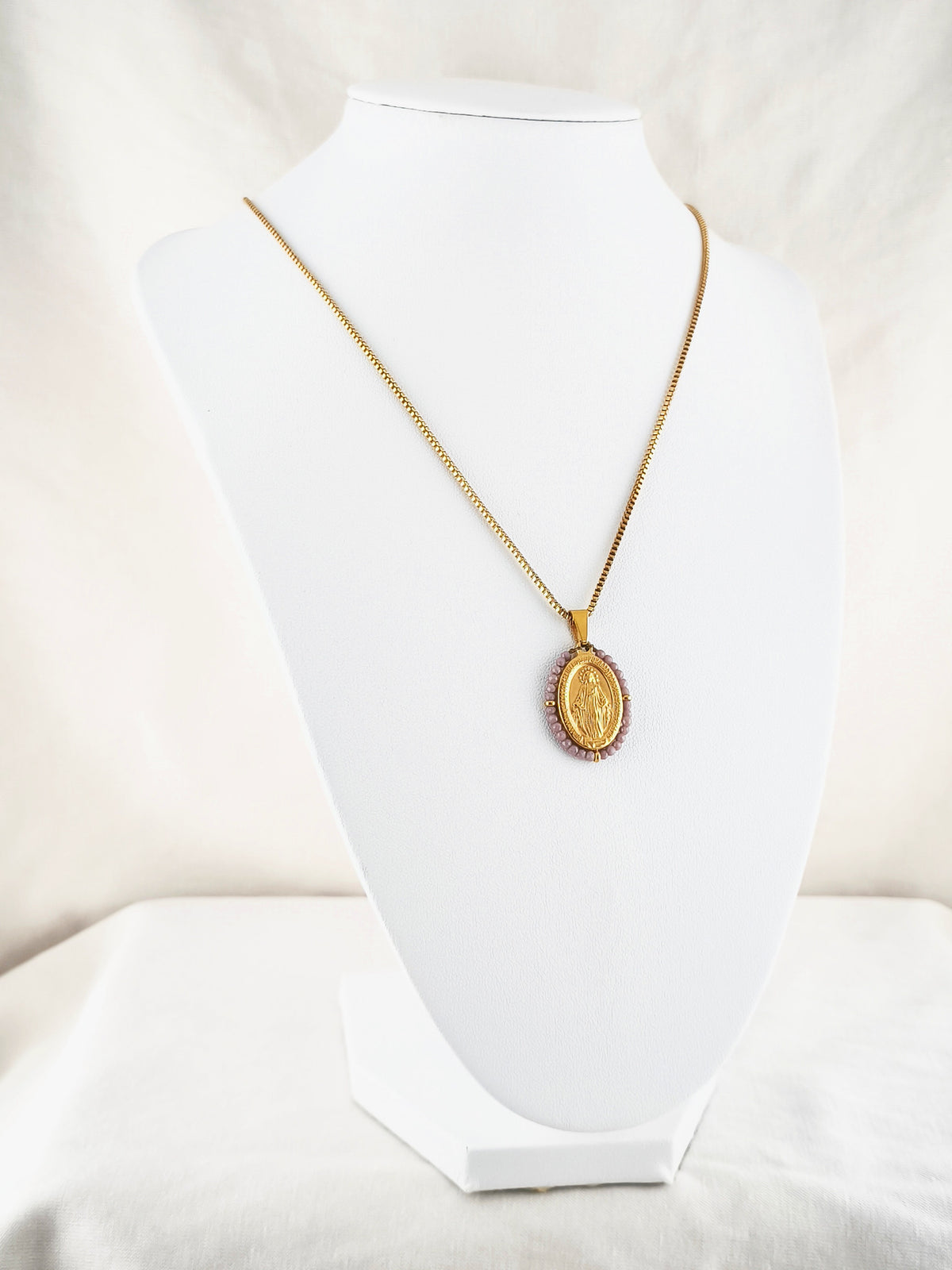 Fio Auréola dourado, com medalha de Nossa Senhora das Graças rodeada de contas lilás, apresentado em expositor.