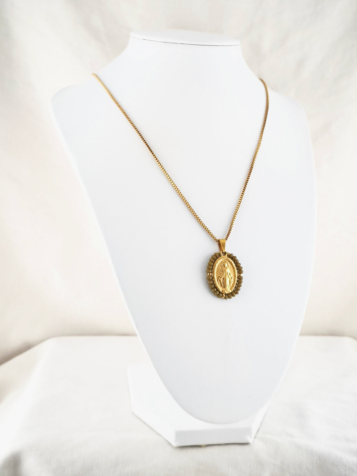 Fio Auréola dourado, com medalha de Nossa Senhora das Graças rodeada de contas caqui, apresentado em expositor.