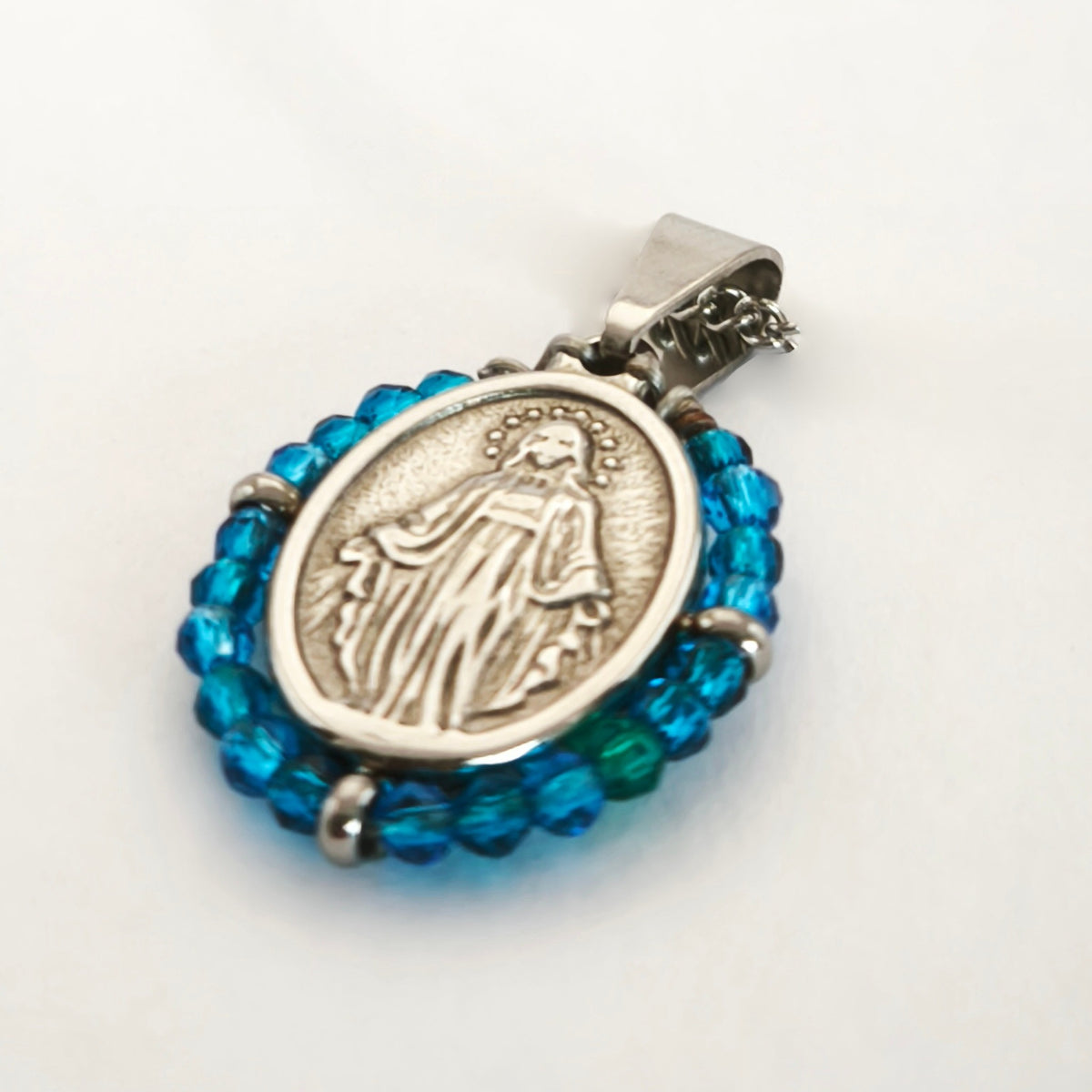 Pendente Orvalho prateado, com medalha de Nossa Senhora das Graças rodeada de contas azul claro, em perspetiva.