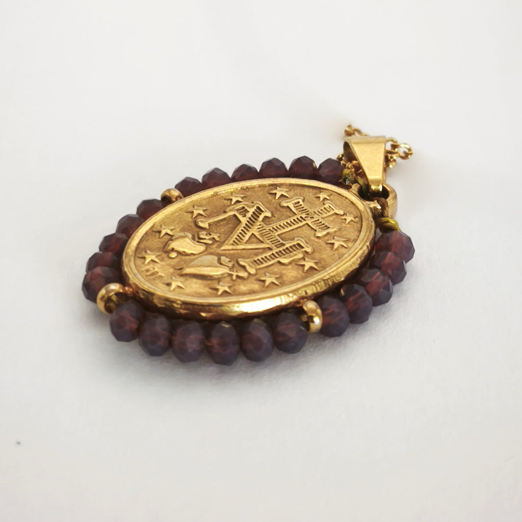 Pendente Auréola dourado, verso da medalha rodeada de contas ameixa, com a letra M, a cruz, os dois corações e as doze estrelas.