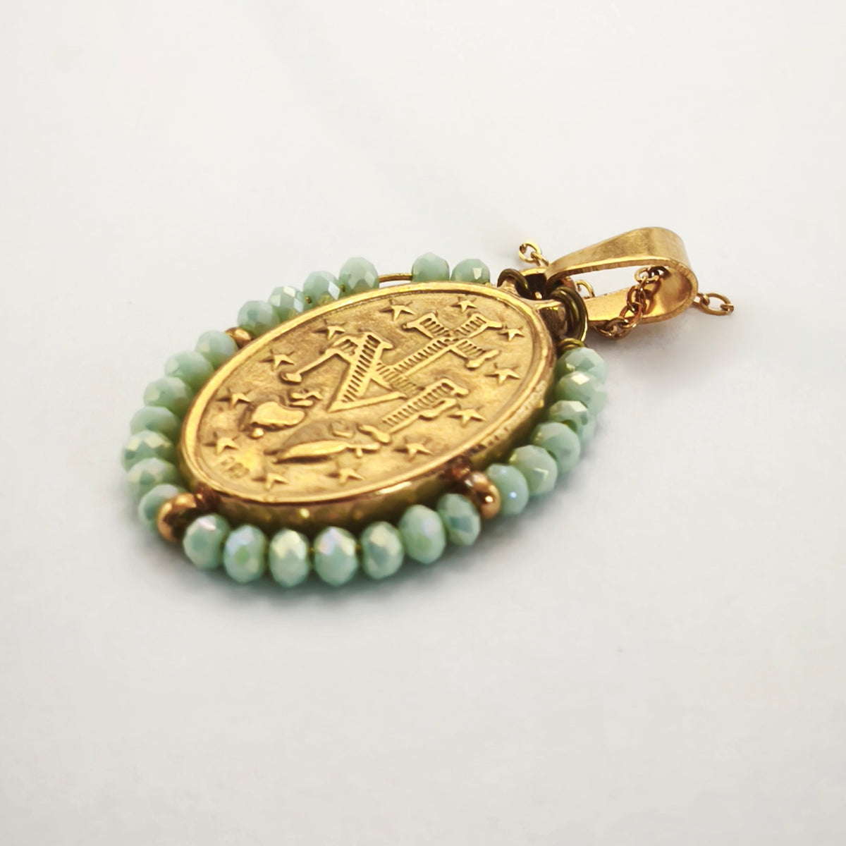 Pendente Auréola dourado, verso da medalha rodeada de contas verde-água, com a letra M, a cruz, os dois corações e as doze estrelas.