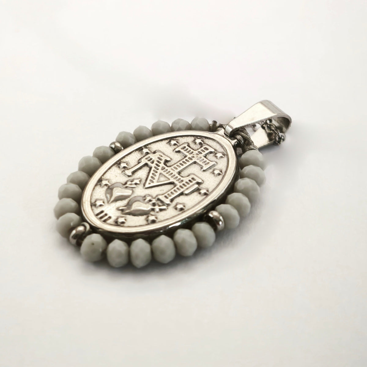 Pendente Auréola prateado, verso da medalha rodeada de contas cinza claro, com a letra M, a cruz, os dois corações e as doze estrelas.